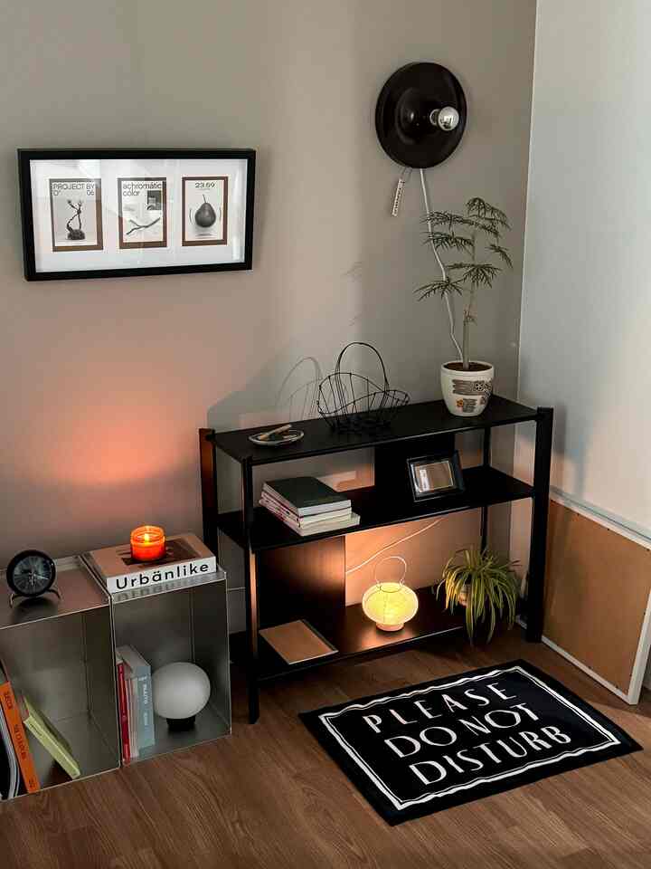 Cozy modern corner space with natural wood floor and white walls, featuring black shelving, framed art, and warm lighting