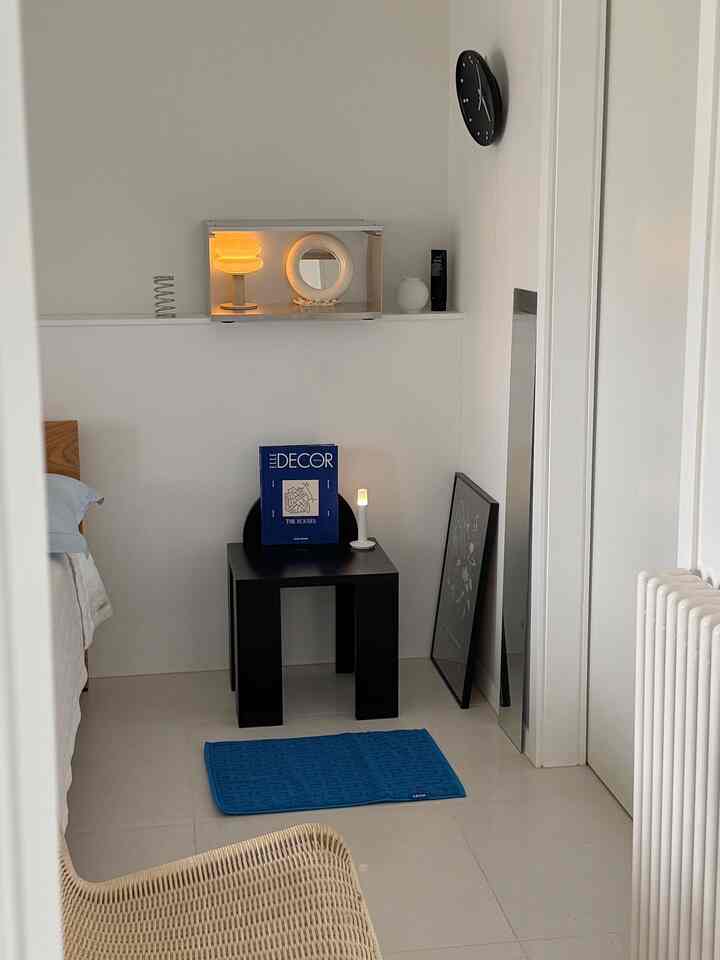 白と黒のトーンにナチュラルウッドを合わせた寝室空間、黒いスツールの上にエルデコの雑誌と照明が置かれたミニマルでモダンなインテリア