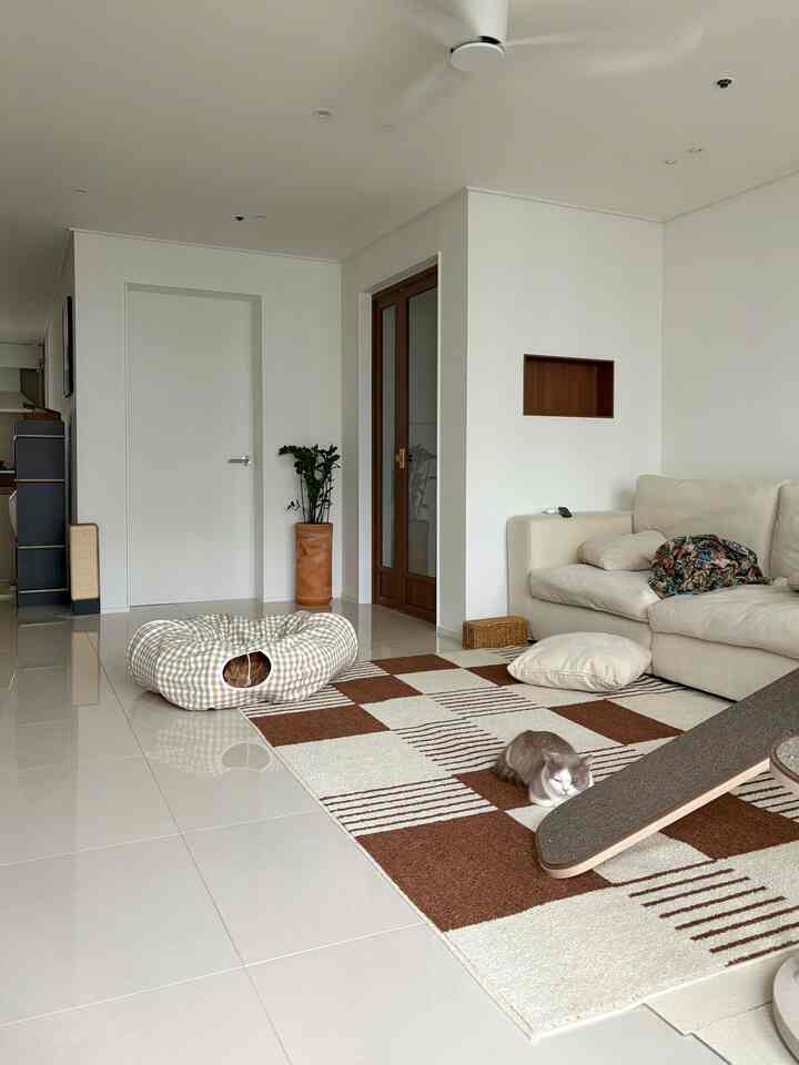 Bright living room with white polished tiles, beige modular sofa, brown checkered rug, cat tower and resting cat