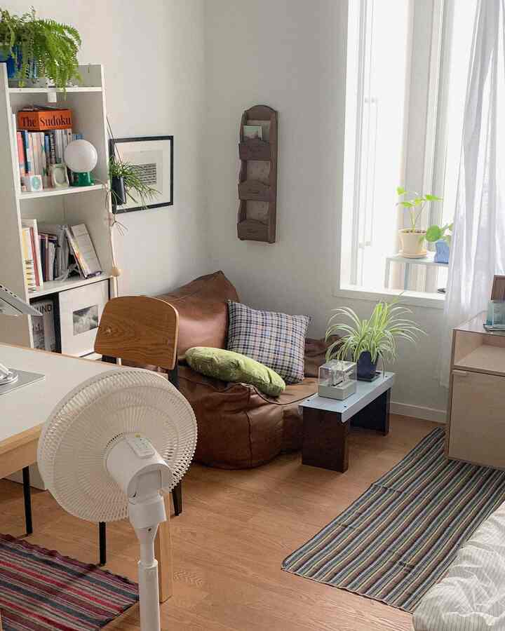 ナチュラルな木目調と白い壁の小さな一人暮らしの部屋、ビーンバッグソファ、本棚、扇風機、植物が配置された雰囲気のあるインテリア