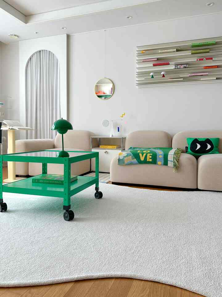 ホワイトトーンの壁と広いラグのあるリビング、ベージュのソファとグリーンのコーヒーテーブルや小物が配置されたモダンでナチュラルな空間