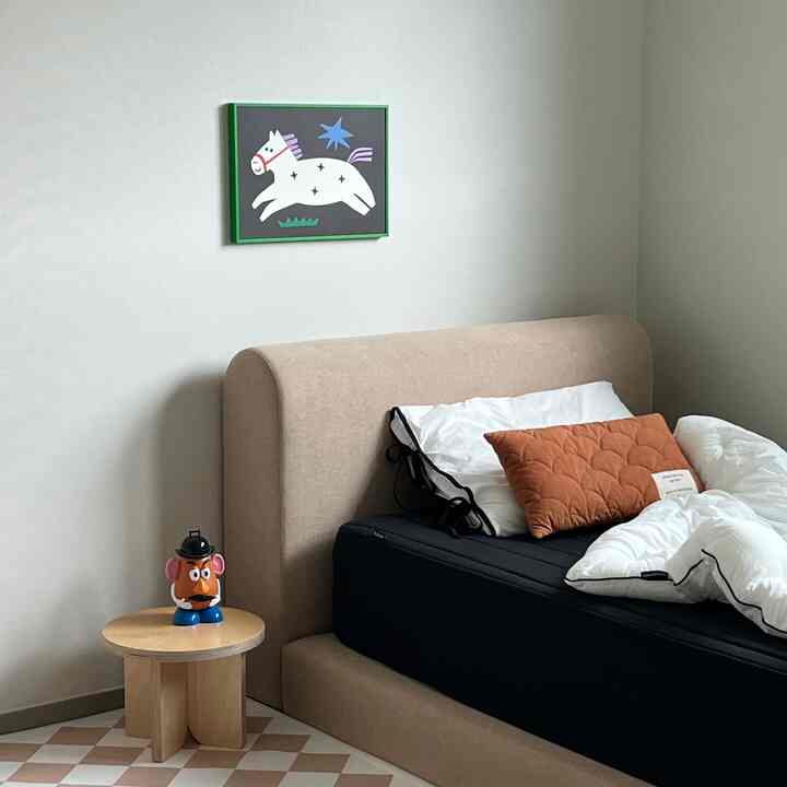 Bedroom with beige bed frame and black mattress, orange pillow on the bed, featuring a cute horse poster on the wall and a round side table