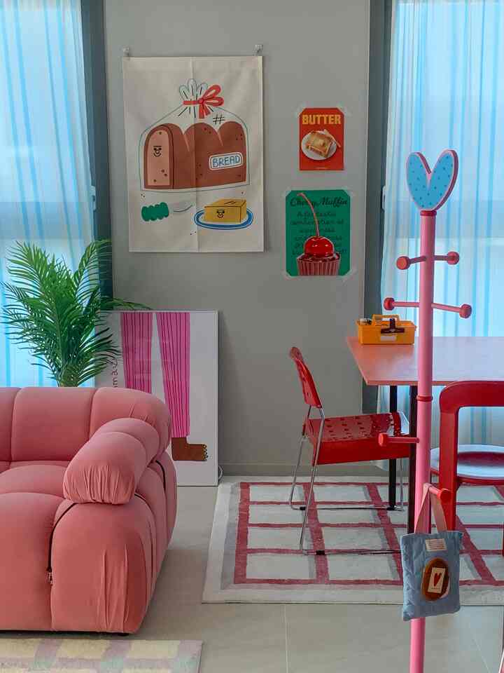 A pink-toned dining room featuring a plush sofa, red dining table with chair, and decorative food posters creating a cozy atmosphere