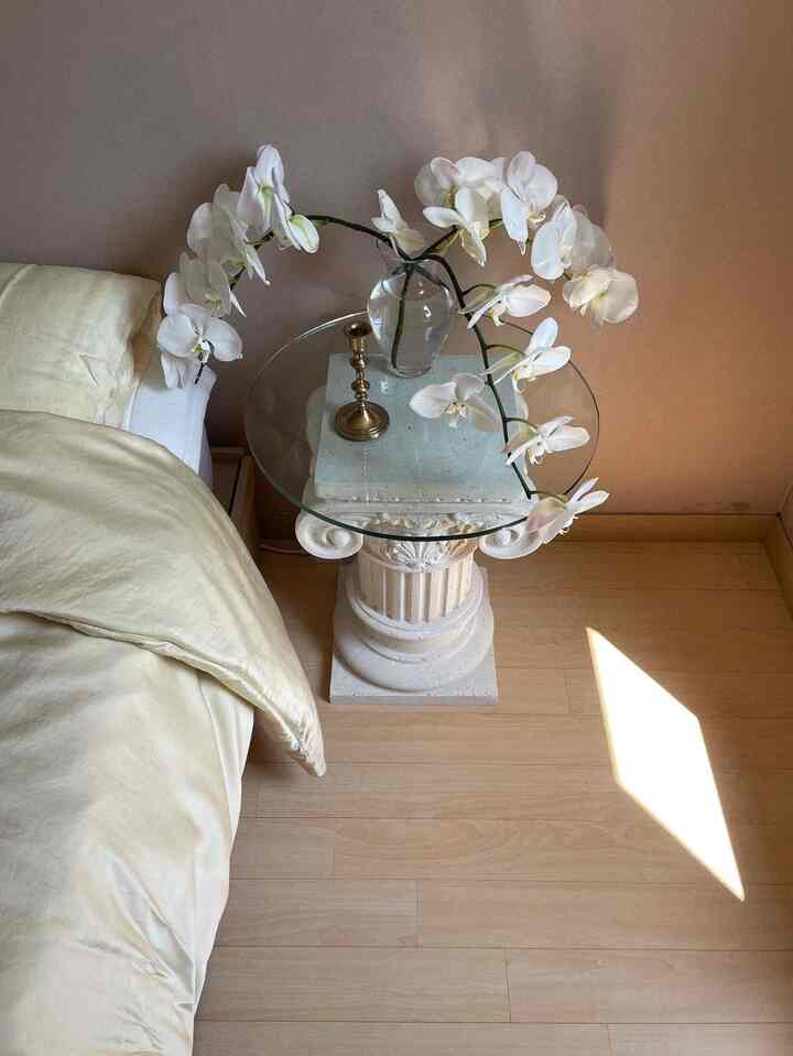 ベージュとホワイトの色調の寝室、アールデコスタイルのナイトテーブルの上に置かれた透明なガラスの花瓶と白い蘭が配置されたシンプルで自然な雰囲気のインテリア