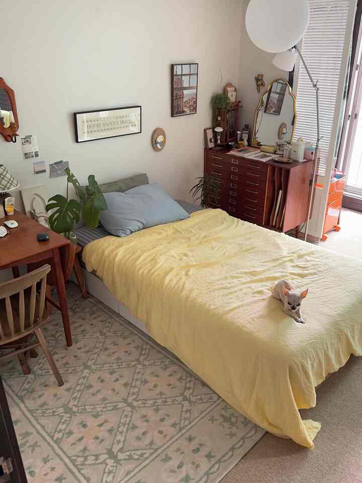 ベージュの壁と黄色い寝具が配置された居心地の良い寝室、木製家具と鏡、ラグが調和した雰囲気のあるインテリア