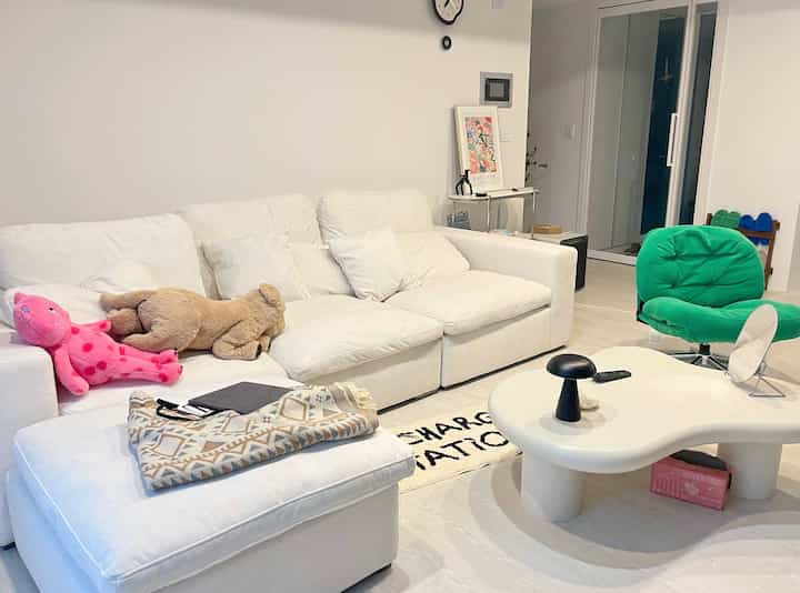 白とベージュの色調のリビング、広いコーナーソファと緑のアームチェア、独特な形のコーヒーテーブルが配置された落ち着いた空間