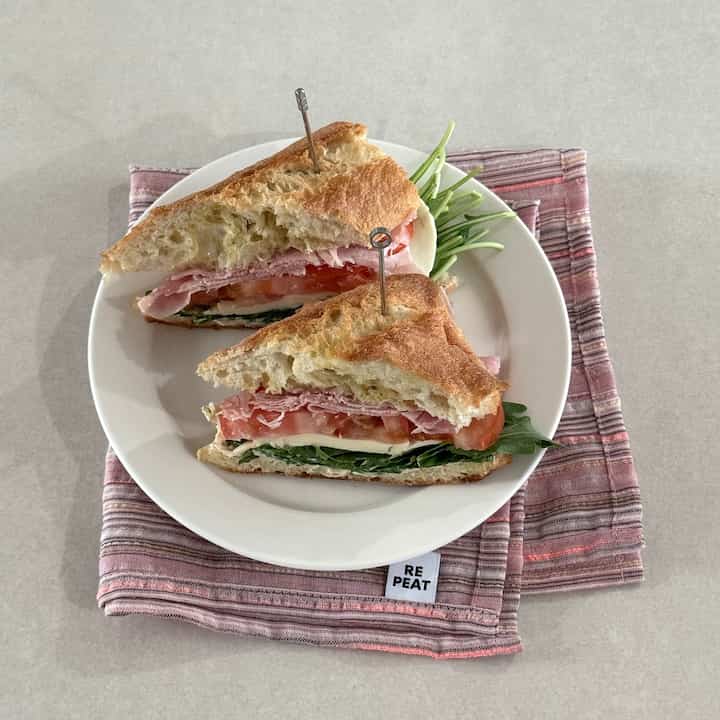 ベージュとホワイトのトーンが調和するテーブルの上にサンドイッチと皿、ピンクのストライプのキッチンクロスが配置されたおうちカフェ風の空間