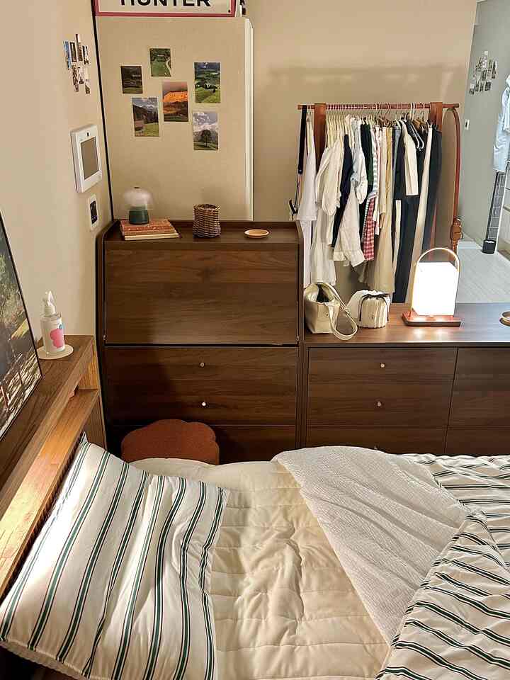 木目調とベージュのワンルームの寝室、ストライプの夏用布団と木製ハンガーラックが配置されたシンプルで落ち着いた空間