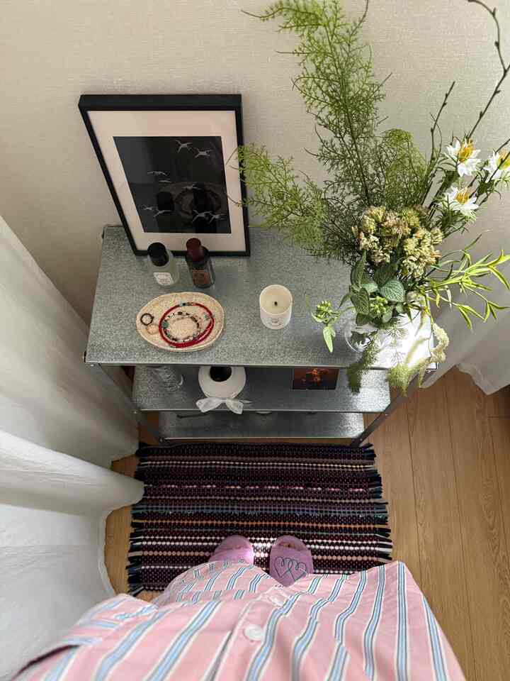 Studio apartment corner with white curtains and wood flooring, featuring an IKEA metal shelf, artificial flowers, artwork, and a rug creating a cozy vibe
