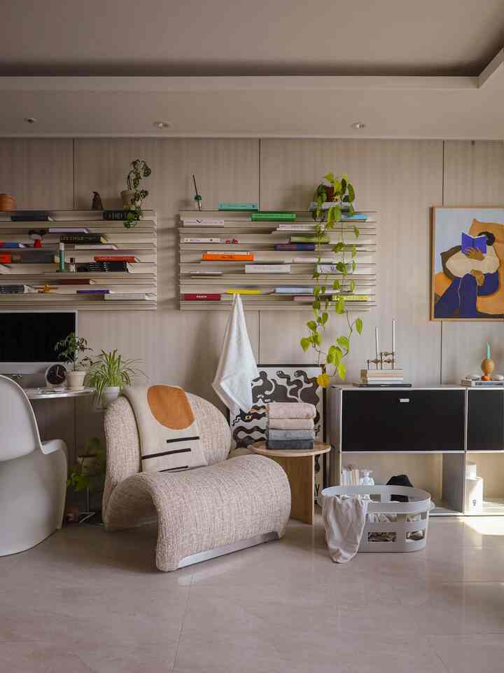 白とベージュの色調のリビング、曲線的なラウンジチェア、棚に本と植物、整理されたタオルが配置された雰囲気のあるインテリア
