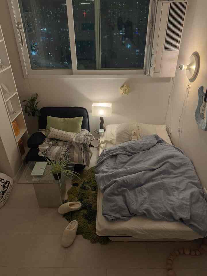 ナチュラルな色調のワンルーム寝室、ベッドとアームチェア、ナイトテーブルの照明が配置された雰囲気のある空間