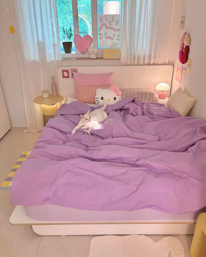 柔らかなパープルの寝具とピンクのクッションが映える小さな寝室、中央のベッドに白い猫がくつろいでいる