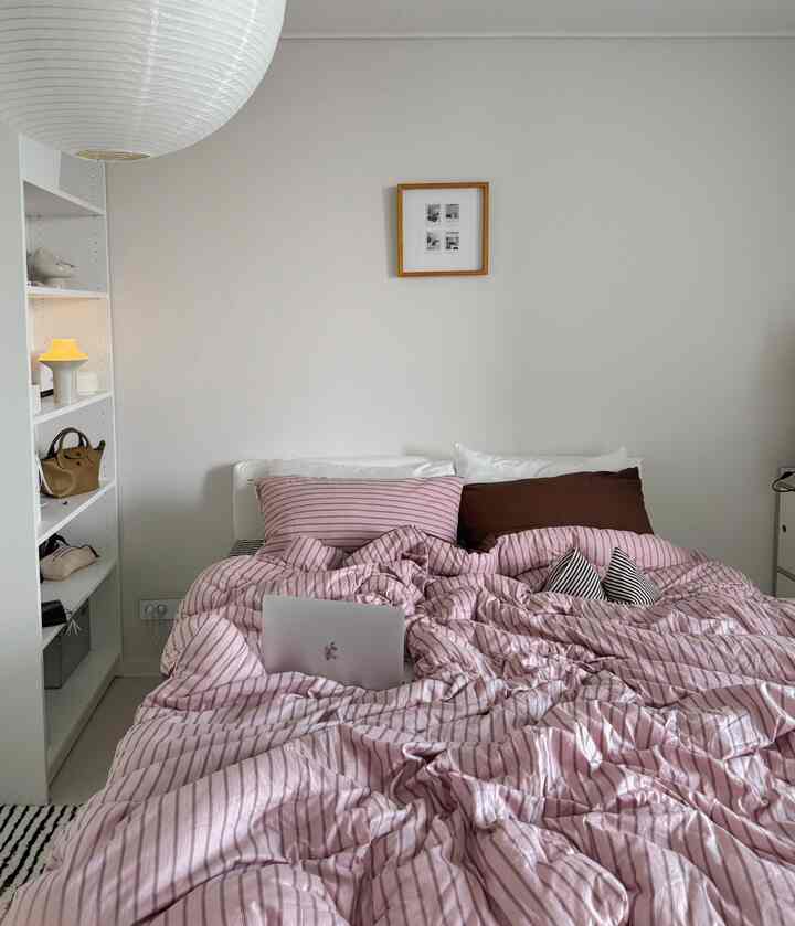 白い壁と棚のあるミニマルな寝室、ピンクのストライプ寝具が印象的な空間