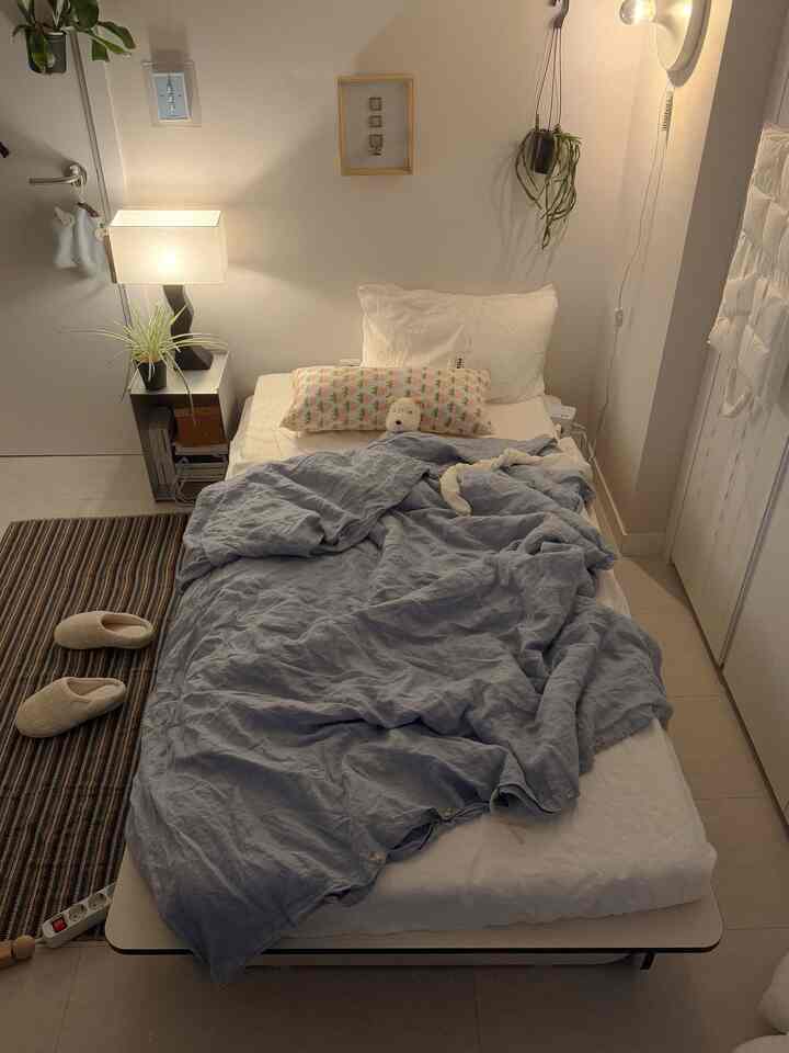 白とベージュのトーンのワンルーム寝室、ヘッドレスベッドとライトグレーの寝具が配置された落ち着いた空間