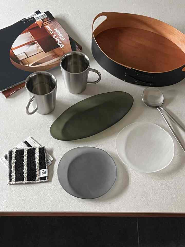 ブラックと木目調が調和したテーブルに、ステンレスのマグカップや様々な皿、ブラックウッドのトレイが配されたミニマルな卓上ディスプレイ