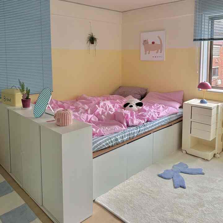 黄色と白の壁が特徴の小さな寝室で、ピンクの寝具と収納家具、植物が配置された温かみのあるインテリア