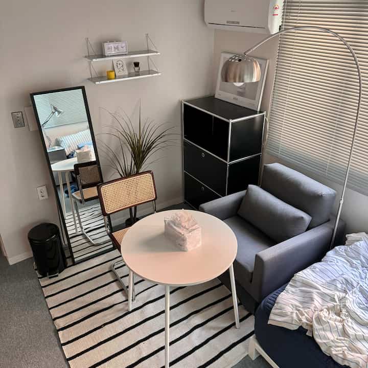 ベージュとグレーの色調の一人暮らし用リビング兼寝室、シンプルでミニマルな木製椅子と丸テーブルが配置された空間