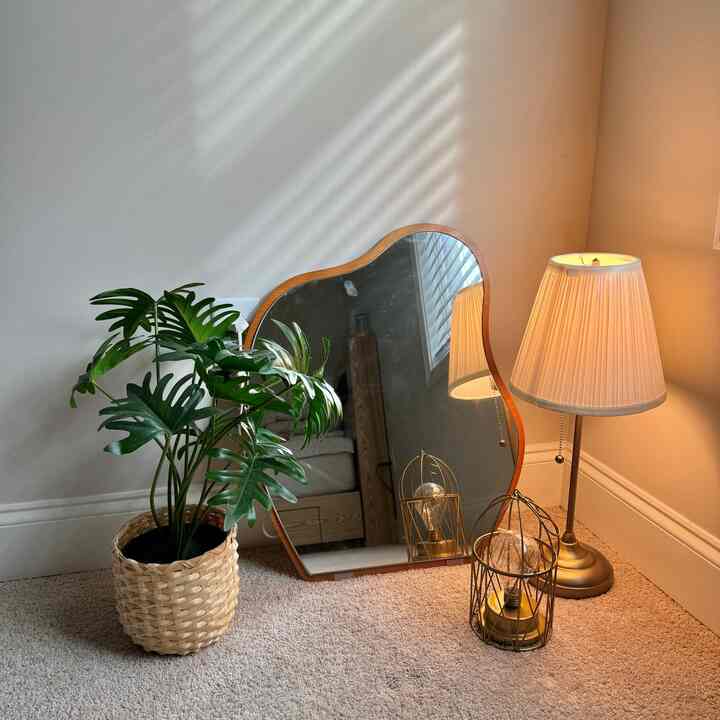 Beige-toned corner featuring a wooden framed mirror, green plant in woven basket, and gold table lamp creating cozy atmosphere