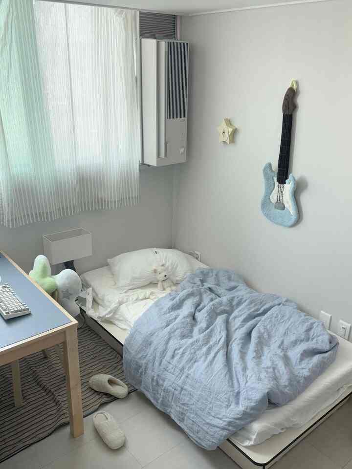 白とナチュラルカラーのコンパクトな寝室、スリムな窓型エアコンとシンプルなベッド・デスクが配置された清潔感のあるインテリア