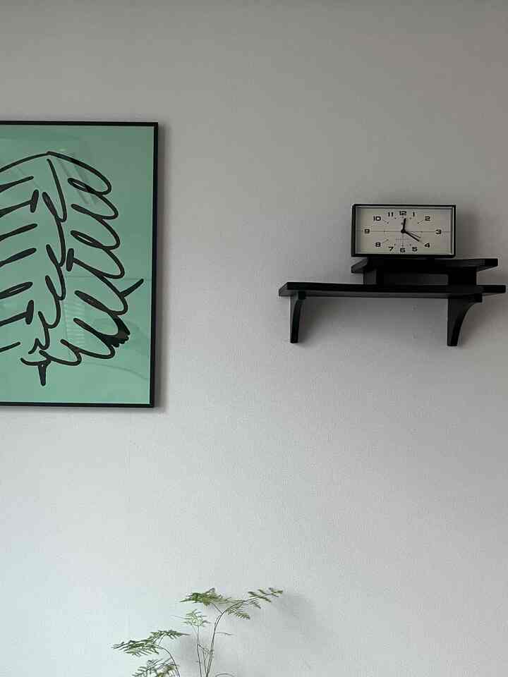 落ち着いた黒の棚と時計、緑の葉のアートが配されたシンプルな白い壁の空間