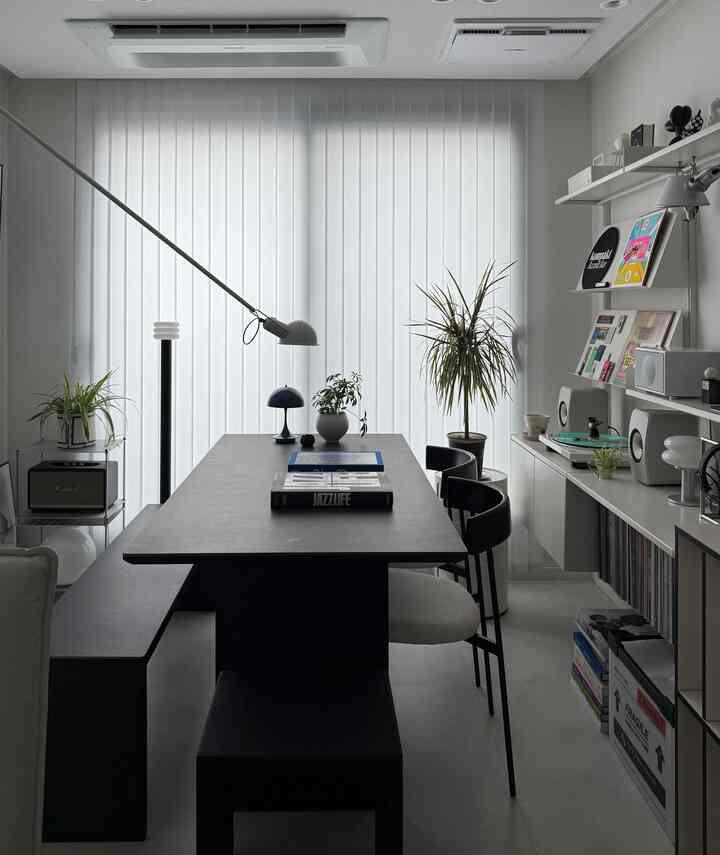 白い縦型ブラインドを背景にしたモダンなダイニングルーム、黒いテーブルと椅子、植物が配置された落ち着いた空間