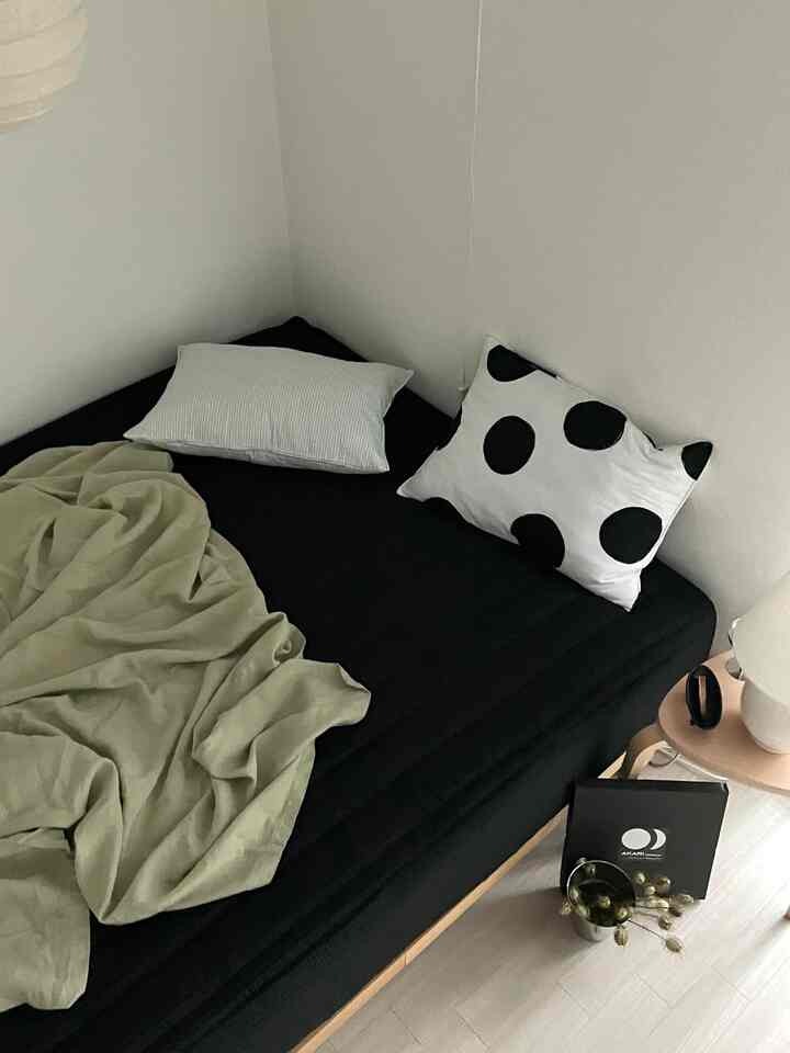 Minimal bedroom corner featuring a black mattress bed with beige blanket, white walls, and a simple wooden side table creating a calm atmosphere