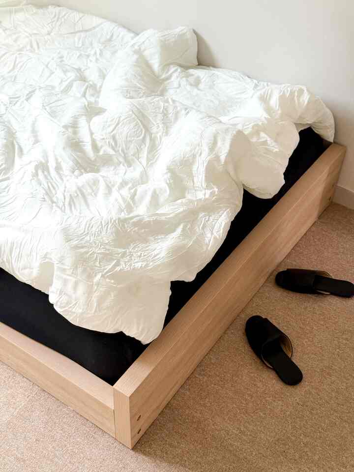 A simple bedroom featuring white bedding and a natural wood bed frame with black indoor slippers placed on beige carpeted floor