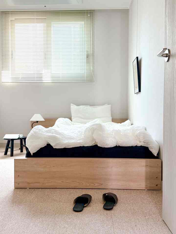 ホワイトと木目調が調和したミニマルな1人用寝室、低い木製ベッドフレームと整った寝具が配置された落ち着いた空間