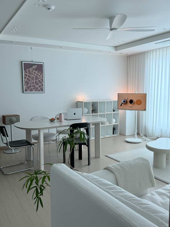 白を基調としたリビング、中央にダイニングテーブルと多様な椅子、天井ファンが配置されたミニマルで清潔感のあるインテリア