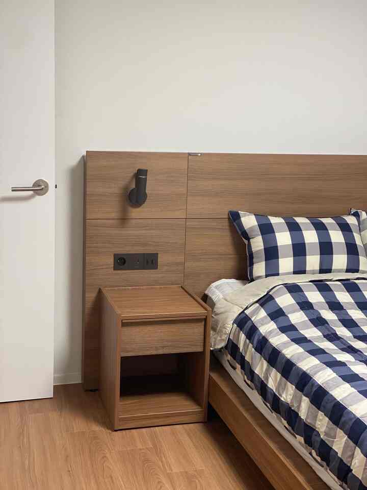 白い壁と木目調のベッド・ナイトテーブルが配置された落ち着いた寝室、青のチェック柄の寝具がアクセント