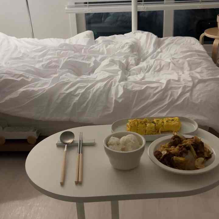 白を基調としたワンルーム寝室、シンプルな簡易テーブルに食事が置かれ、暖かみのある印象的な空間