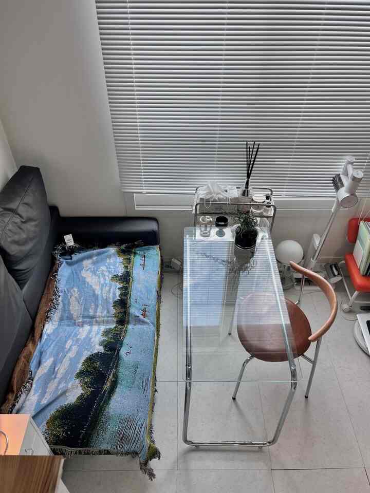 白を基調としたワンルームのリビング、ブラインド付きの窓と黒いソファ、ガラステーブルが配置された清潔感あるナチュラルモダンなインテリア