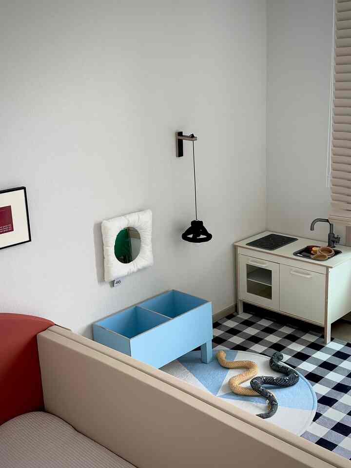 ホワイトとブルーの色調の子供部屋、幼児用バンパーベッドとキッズ用キッチン遊びセット、壁付けライトが配置された可愛らしい空間