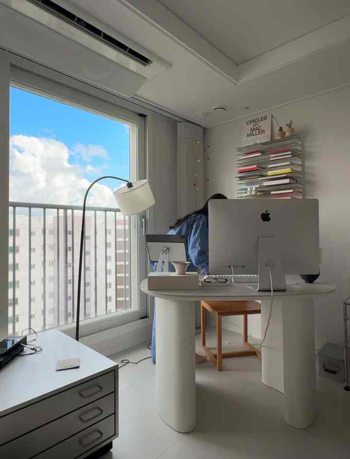 ホワイトトーンのアパートのホームオフィス、広いデスクにiMacとフロアランプが配置された雰囲気のある空間。