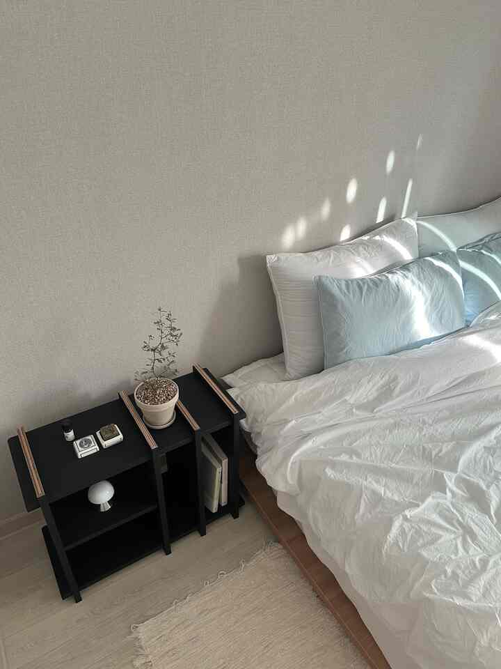 Bright white and natural tone bedroom featuring wooden bed frame with white bedding on right, black side table with plant on left in a clean modern setting