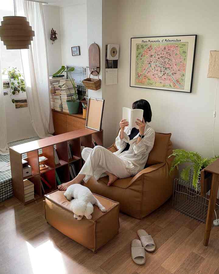 ナチュラルな木目調とブラウンレザー家具が配置された居心地の良い狭い部屋、ビーズクッションソファとスツールの上で犬が休む読書スペース
