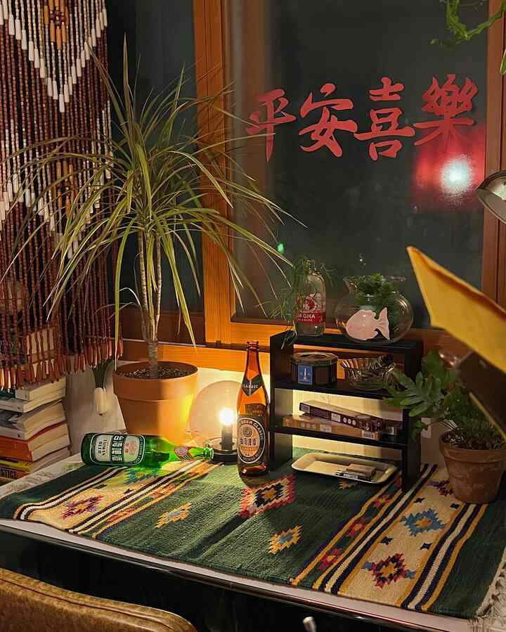 緑色とダークブラウンのトーンのデスク上にインド製キリムラグ、鉢植え、書籍、ヴィンテージ瓶、テーブルランプが配置された雰囲気のあるインテリア