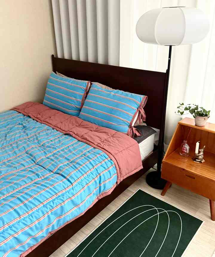 A bedroom with brown wooden bed frame and blue striped bedding, featuring a green rug, wooden nightstand, and white floor lamp creating a modern, natural atmosphere
