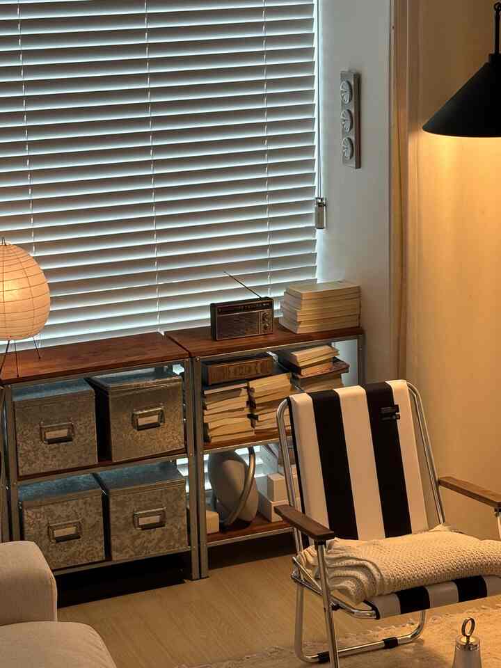 ホワイトブラインドと木目調の棚、ストライプのアームチェアが配置されたモダンなリビングスペース