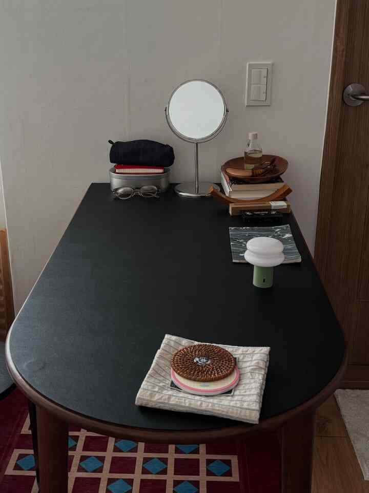 黒いテーブルトップと木目調の脚が見える狭い部屋の簡易化粧台、丸い鏡や本が配置された落ち着いた空間