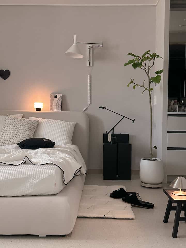 A modern bedroom in white and light gray tones featuring black accent cushions and layered lighting creating a clean atmosphere