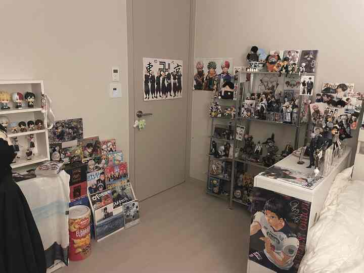 白と茶色のトーンの狭いオタク部屋、フィギュアと漫画雑誌が棚にぎっしり並べられたファンの空間