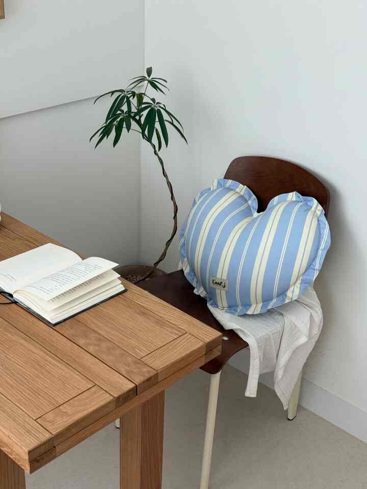 白い壁と木目調の机と椅子が配置されたコーナーのホームオフィス空間、ハート形クッションと植物がアクセントの落ち着いた雰囲気