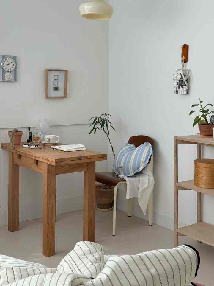 白い壁と木目調家具が調和する寝室で、木製のデスクとパステルのクッションが置かれた椅子、観葉植物が配された落ち着いた雰囲気のインテリア