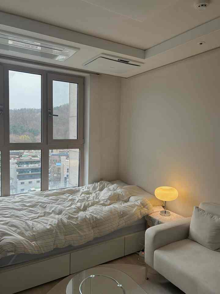 白とナチュラルカラーの6坪オフィステル寝室、ベッドとソファ、ナイトテーブルランプが配置された雰囲気のあるインテリア