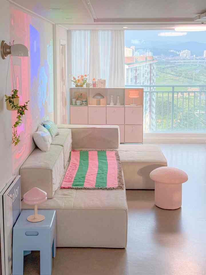 Modern living room in white and pink tones featuring a sectional sofa, pink stool, sheer curtains, and wall sconces creating a cozy atmosphere