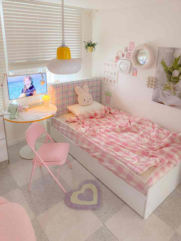 Pastel-toned compact bedroom featuring a pink checkered bed, pink folding chair, cute cushion, and a yellow table lamp creating cozy atmosphere