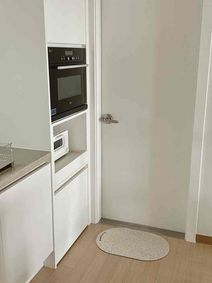 ホワイトとベージュのトーンの狭いキッチンスペース、ビルトインのオーブンとトースター、入り口前に配置されたオセルピックのバスマットが印象的な清潔感のあるインテリア