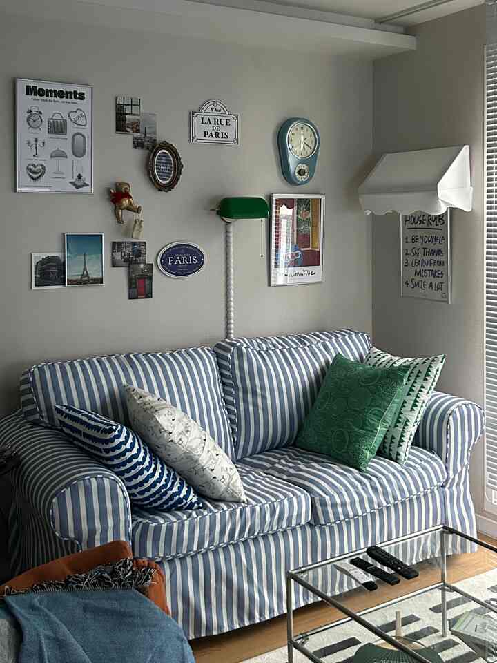 ホワイトトーンのリビングにブルーとホワイトのストライプ布張りソファを中心に配置し、ヴィンテージの額縁と多彩なクッションが調和した温かみのある空間