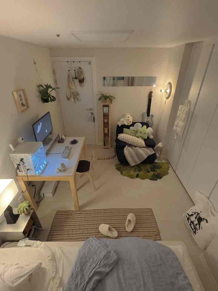 明るいベージュトーンのワンルーム、ベッドとデスク、アルテックチェア、壁ブラケットライト、植物が配置された雰囲気のあるモダンなインテリア